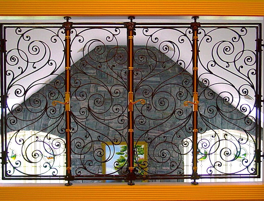 Dark scrolled window guard seen from indoors, brass handles and stained glass beyond